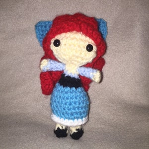 Puede incluir: Una mu&ntilde;eca de crochet con pelo rojo, un vestido azul y zapatos negros. La mu&ntilde;eca tiene una cara blanca con ojos negros.