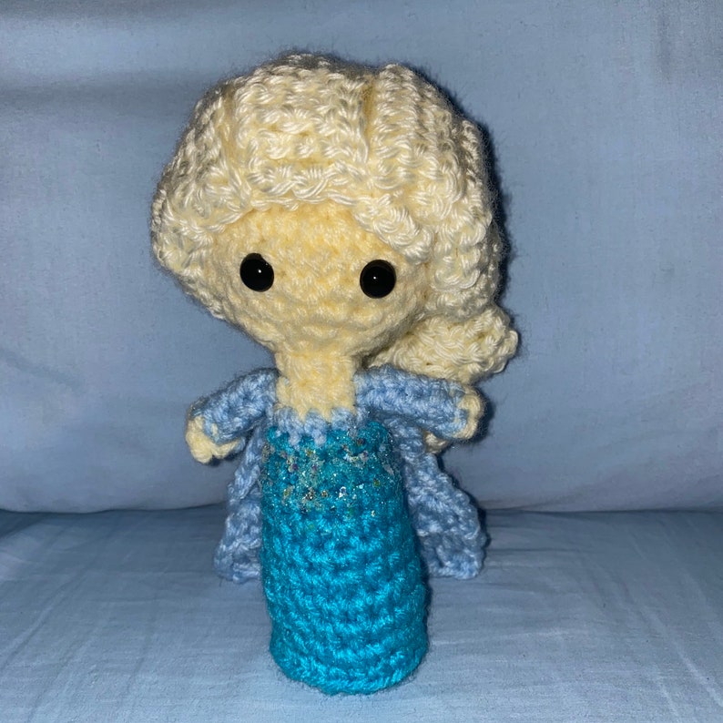 May include: A crocheted doll with blonde hair and a blue dress. The doll has black eyes and a light yellow face.