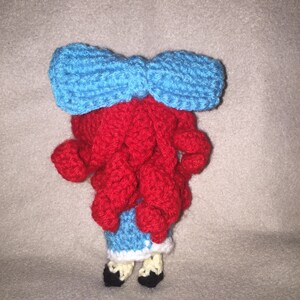 Puede incluir: Una mu&ntilde;eca de crochet con pelo rojo, un lazo azul y un vestido azul y blanco.