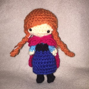 May include: Crocheted doll with red hair, a blue dress, and a pink cape. The doll has a white face with black eyes and a black mouth.
