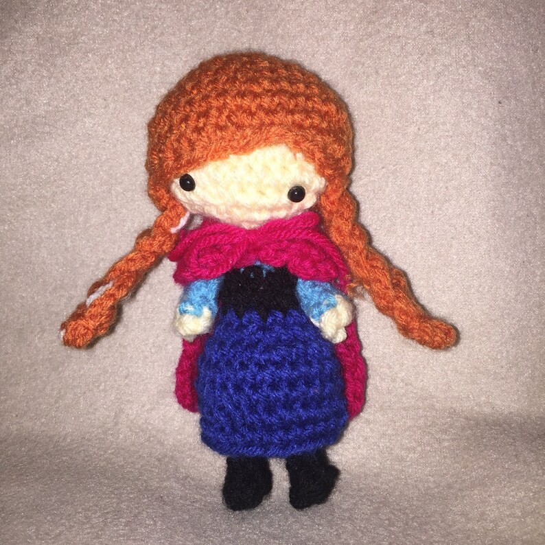 May include: Crocheted doll with red hair, a blue dress, and a pink cape. The doll has a white face with black eyes and a black mouth.