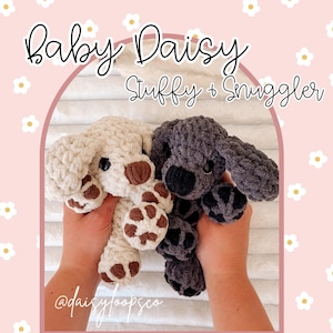 May include: Two crocheted dog stuffed animals, one cream and one gray, are held up against a pink background. The cream dog has brown accents on its paws and ears. The gray dog has black accents. The text "Baby Daisy Stuffy + Snuggler" is at the top.
