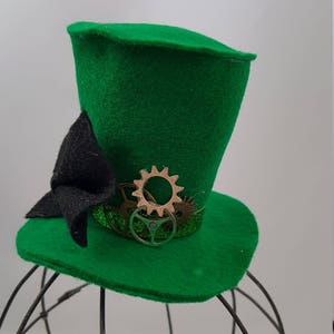 May include: A green felt top hat with a black bow and gold and silver gears. The hat is sitting on a wire hat stand.