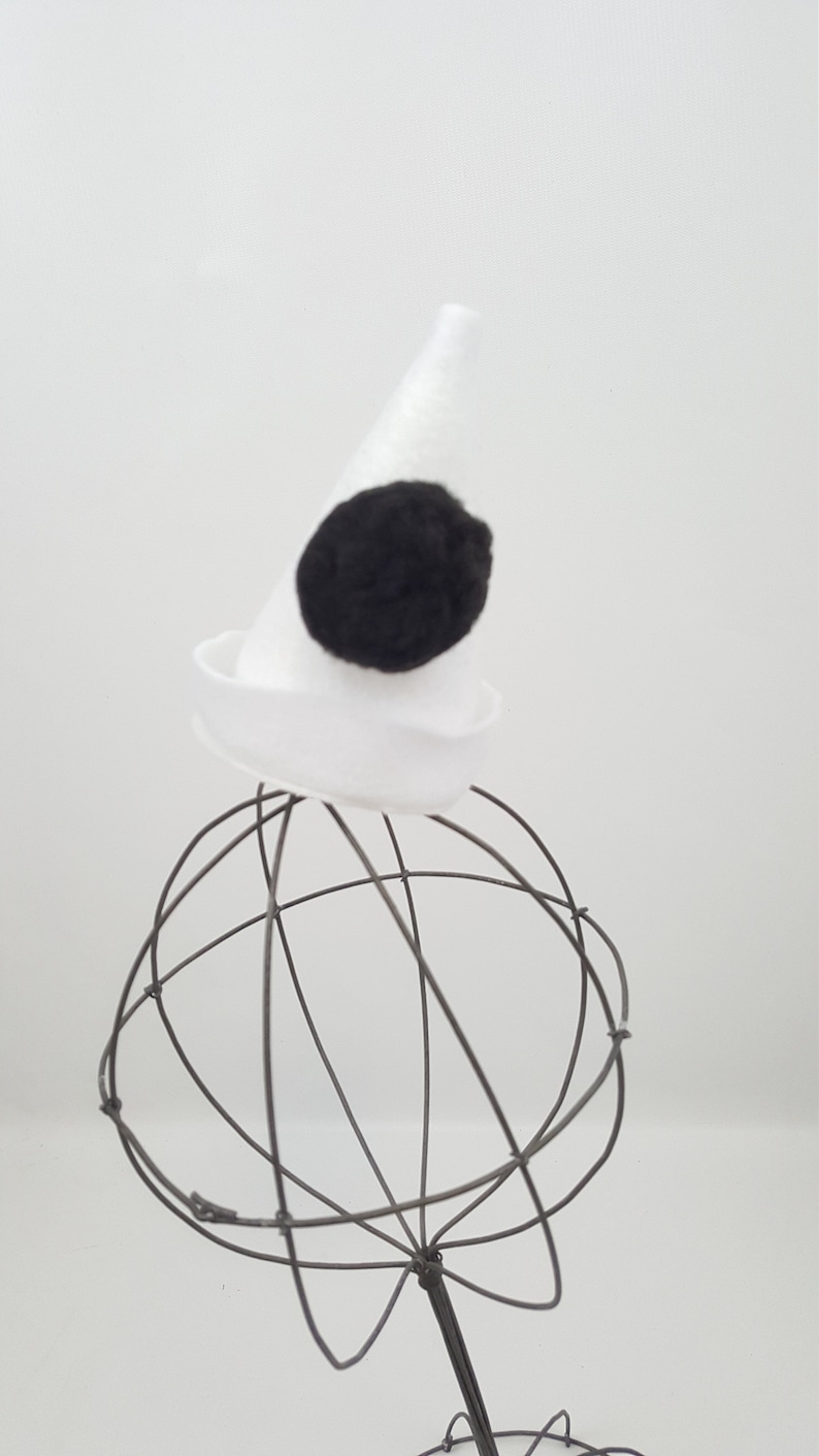 May include: A white felt cone hat with a black felt pom-pom on top. The hat is sitting on a wire hat stand.
