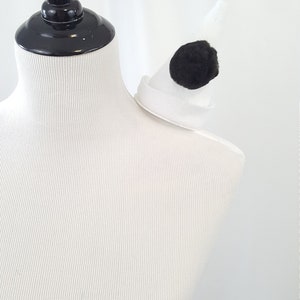 May include: A white and black costume accessory with a white felt base and a black pom-pom. The accessory is designed to be worn on the shoulder.