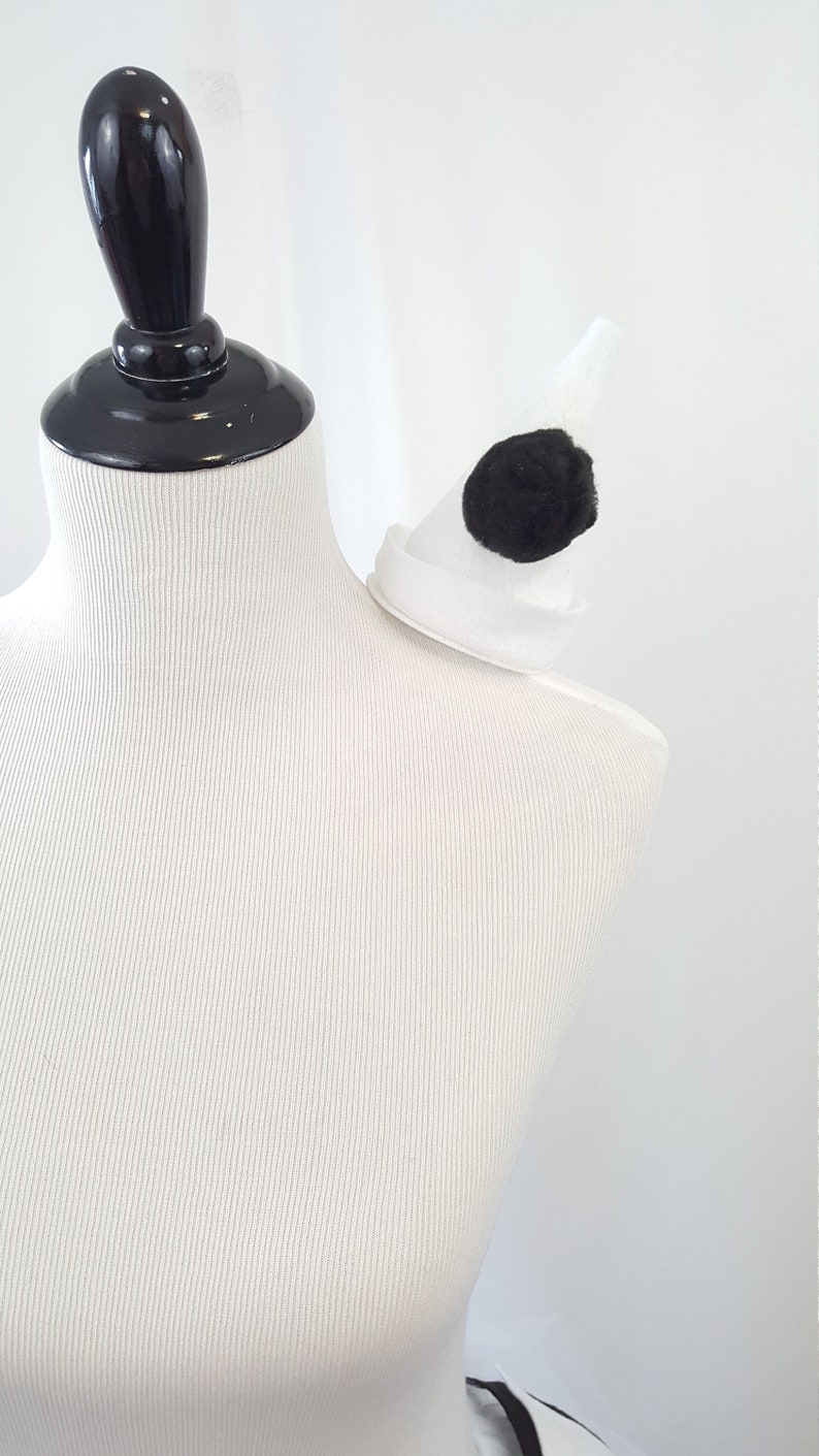 May include: A white and black costume accessory with a white felt base and a black pom-pom. The accessory is designed to be worn on the shoulder.