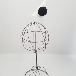 May include: A white wire hat stand with a white cone hat with a black pom-pom on top.