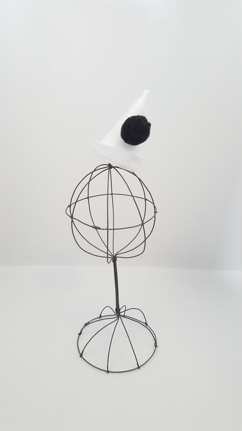 May include: A white wire hat stand with a white cone hat with a black pom-pom on top.