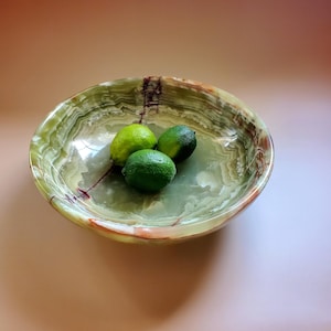 May include: A green onyx bowl with a swirling pattern, holding three limes. The bowl has a polished finish and a natural, organic aesthetic. The limes are various shades of green.
