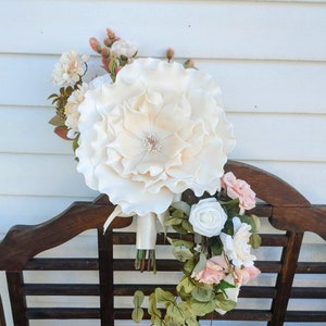May include: A white and blush bridal bouquet with a large, ruffled white flower, greenery, and smaller white and blush flowers.