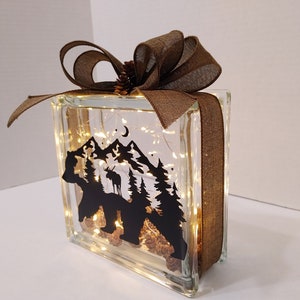 May include: A glass block featuring a silhouette of a bear, mountains, and trees. The block is illuminated with fairy lights and tied with a brown ribbon.