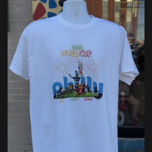 May include: A white short-sleeve t-shirt featuring a 2026 World Cup graphic. The design includes the text '2026 WORLD CUP' above a central illustration of a soccer trophy, fireworks, and the word 'philly' with cartoon mascots and 'USA MEXICO CANADA'.