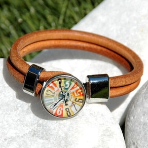 Puede incluir: Un brazalete de cuero marrón con un cierre plateado y un dial redondo colorido con números.
