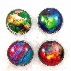 Puede incluir: Cuatro botones redondos con borde plateado y diseños abstractos coloridos. Cada botón presenta un patrón de remolino diferente en azul, verde, rojo, amarillo y naranja.
