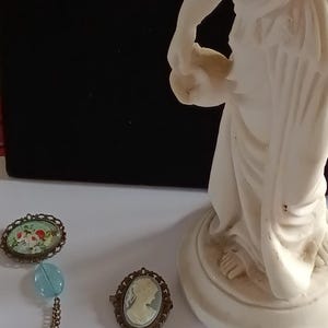 Puede incluir: Broche de estilo vintage con dos colgantes ovalados, uno con un diseño floral y otro con un camafeo. Ambos colgantes están conectados por una doble cadena y adornados con cuentas azul claro. Una estatua blanca está en el fondo.