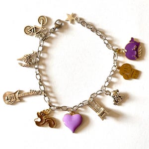 Puede incluir: Pulsera de abalorios plateada con varios dijes, incluyendo una bicicleta, guitarra, estrella, corazón, tetera y osito de peluche. El dije de corazón es lavanda. Fondo blanco.