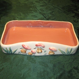 May include: A rectangular ceramic dish with a pink interior and a white exterior. The dish is decorated with a green leafy design and a row of painted mushrooms with pink caps and brown stems.