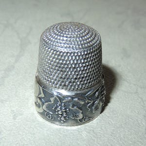 Könnte beinhalten: Ein silberner Fingerhut mit strukturierter, geriffelter Oberseite und einem dekorativen Band. Das Band zeigt ein Weinreben-Design mit Blättern und Trauben. Der Fingerhut ist ein Nähwerkzeug.