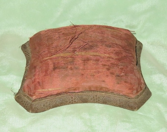 May include: A vintage, square-shaped pincushion with a red fabric top and a dark brown wooden base. The fabric is worn and has visible threads.