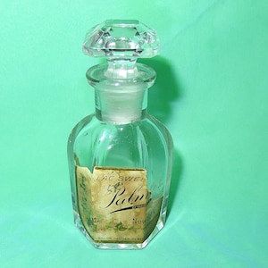 May include: Clear glass perfume bottle with a faceted stopper. The bottle has a label that reads "LAC SWEET Palm" with additional text. The bottle is empty and sits on a green background.