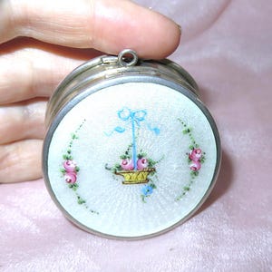 May include: A small, round, silver trinket box with a hinged lid. The lid has a white enamel surface decorated with a floral design of pink roses, a yellow basket, and blue accents. The box has a small loop for a chain.