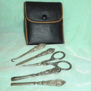 Op de afbeelding: Een zwarte kunstlederen vouwetui met beige rand en drukknoopsluiting, vergezeld van vier zilverkleurige metalen verzorgingsinstrumenten. Elk instrument heeft een versierd handvat met bloemmotief, waaronder een knopenhaak, een nagelvijl, een schaar en een nagelriemduwer.