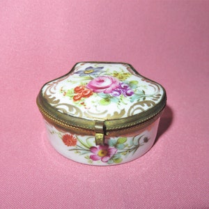 Op de afbeelding: Een klein, wit porseleinen doosje met een bloemenmotief en gouden accenten. Het doosje heeft een scharnierend deksel met een messing sluiting.