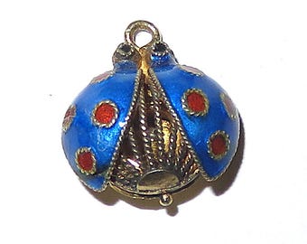 May include: A small, three-dimensional ladybug charm featuring a bright blue enamel finish with red circular spots. The body is crafted from gold-toned metal with a textured, filigree-style underbelly and a small loop at the top for attachment.