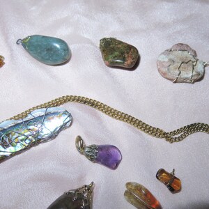 May include: A collection of gemstone pendants, including a green stone, a pink and white stone, a purple stone, and a shell wrapped in wire. The pendants are on a gold chain.