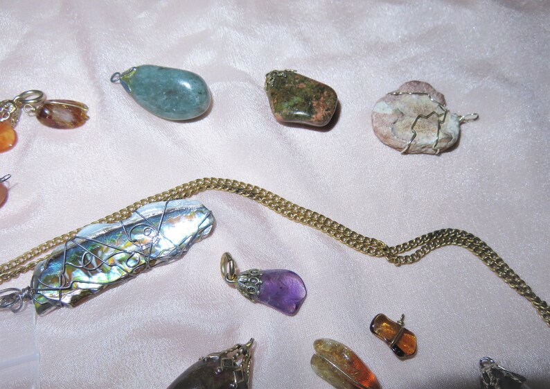 May include: A collection of gemstone pendants, including a green stone, a pink and white stone, a purple stone, and a shell wrapped in wire. The pendants are on a gold chain.