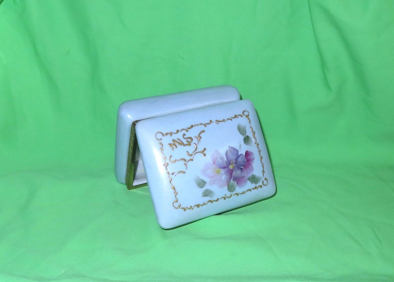 May include: A white porcelain trinket box with a gold trim and floral design. The lid features a delicate floral design with purple and pink flowers.