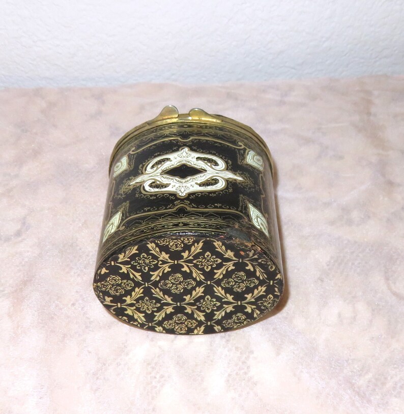 May include: A black cylindrical box with a gold and white floral design. The box has a hinged lid and a brass clasp.
