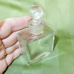 May include: A clear glass bottle with a round glass stopper. The bottle is square shaped with slightly rounded edges.