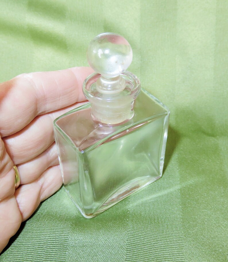 May include: A clear glass bottle with a round glass stopper. The bottle is square shaped with slightly rounded edges.