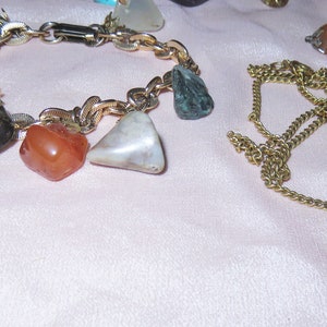 May include: Two gold chains with gemstone pendants. One chain has a bracelet with multiple stones, including red, green, and white. The other chain has a single, large, white and brown stone pendant.