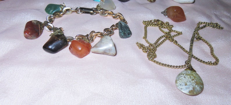 May include: Two gold chains with gemstone pendants. One chain has a bracelet with multiple stones, including red, green, and white. The other chain has a single, large, white and brown stone pendant.