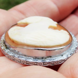 May include: A cameo brooch with a white cameo of a bird design set in a silver tone metal frame with a detailed floral pattern.