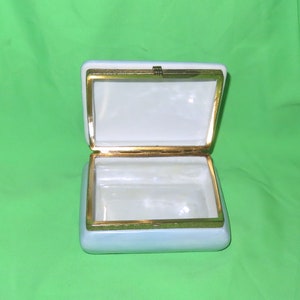 May include: An empty white porcelain trinket box with a gold trim and a hinged lid. The box is open and the inside is white.
