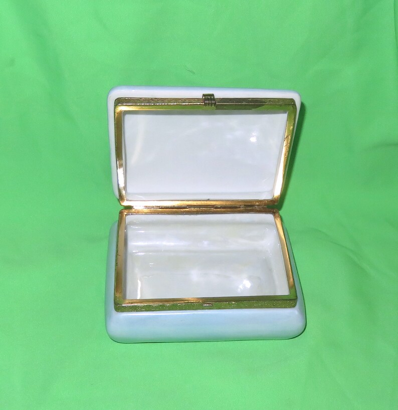 May include: An empty white porcelain trinket box with a gold trim and a hinged lid. The box is open and the inside is white.