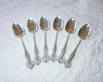 May include: Six silver spoons with ornate handles. The spoons are arranged in a semi-circle on a white surface.