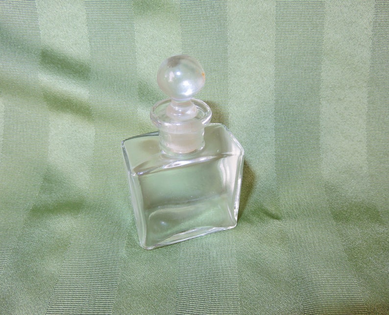 May include: A clear glass bottle with a round glass stopper. The bottle is square shaped and has a smooth surface.