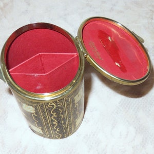 May include: A small, round, black and gold trinket box with a red velvet interior. The box has a hinged lid and is divided into three compartments.