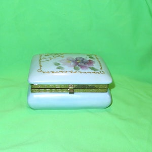 May include: A light blue porcelain trinket box with a hinged lid. The lid features a floral design with pink and purple flowers and gold accents.