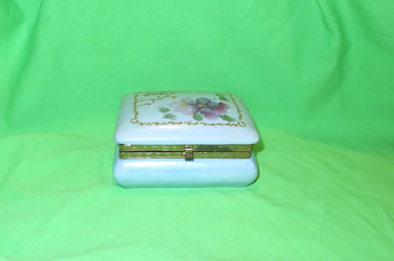 May include: A light blue porcelain trinket box with a hinged lid. The lid features a floral design with pink and purple flowers and gold accents.