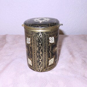 May include: A black cylindrical container with a gold lid and intricate gold and white designs. The container has a hinged lid with a small brass handle.