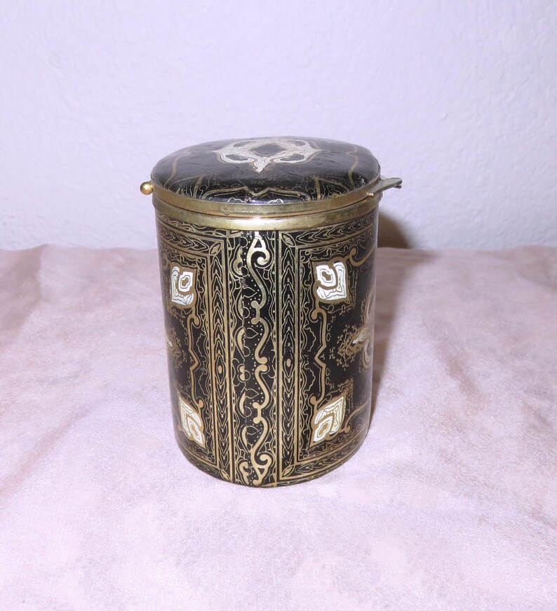 May include: A black cylindrical container with a gold lid and intricate gold and white designs. The container has a hinged lid with a small brass handle.