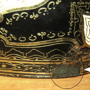 May include: A black and gold decorative object with intricate designs and a chipped area on the bottom edge. The object is made of metal and has a circular shape.