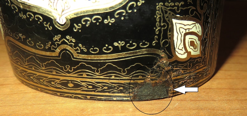 May include: A black and gold decorative object with intricate designs and a chipped area on the bottom edge. The object is made of metal and has a circular shape.