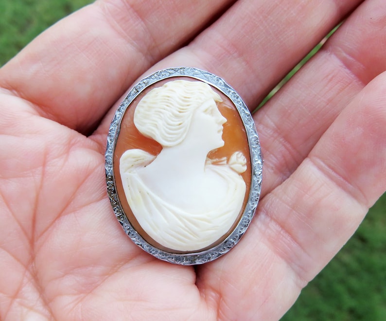 May include: A cameo brooch featuring a carved portrait of a woman with a flower in her hair, set in a silver oval frame with a textured edge.