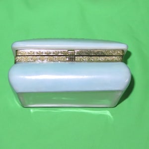 May include: A white porcelain trinket box with a gold-colored hinged lid. The box has a rectangular shape with rounded edges.